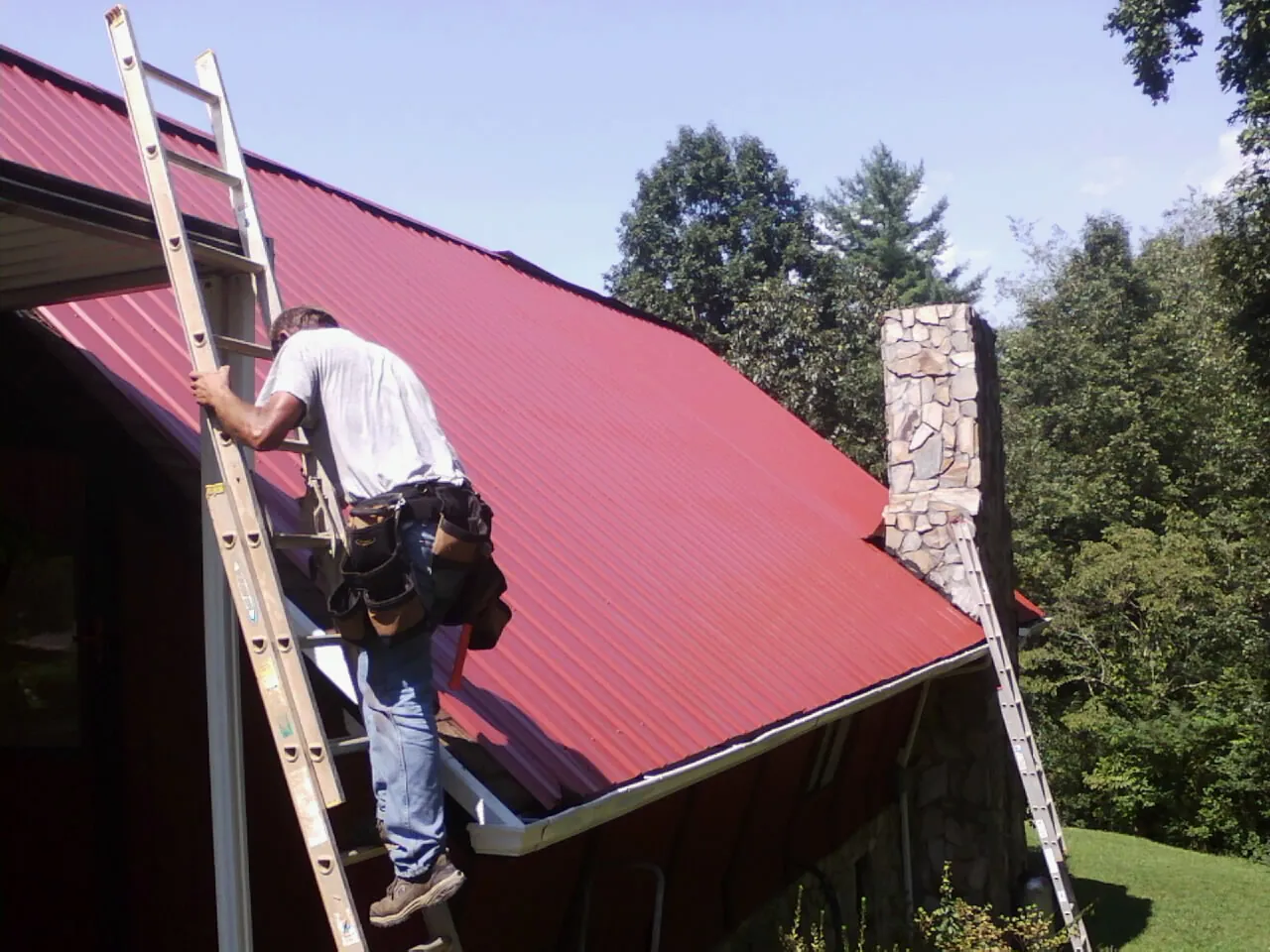 Roofer on a ladder beside a red metal roof and stone chimney — Soffit & Fascia Repair by Roofing Brothers in Renner
